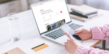 Pessoa utilizando celular e laptop em Airbnb - Créditos: depositphotos.com / VitalikRadko