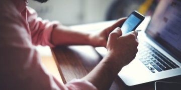 Homem utilizando celular e laptop – Créditos: Créditos: depositphotos.com / GaudiLab