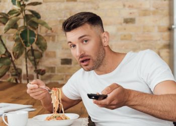 Homem assistindo TV e almoçando - Créditos: depositphotos.com / IgorVetushko